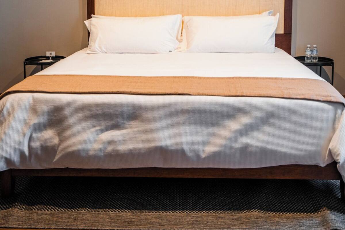 Una cama bien hecha con cabecera de madera, dos mesitas de noche con botellas de agua y dos luces fijadas a la pared.