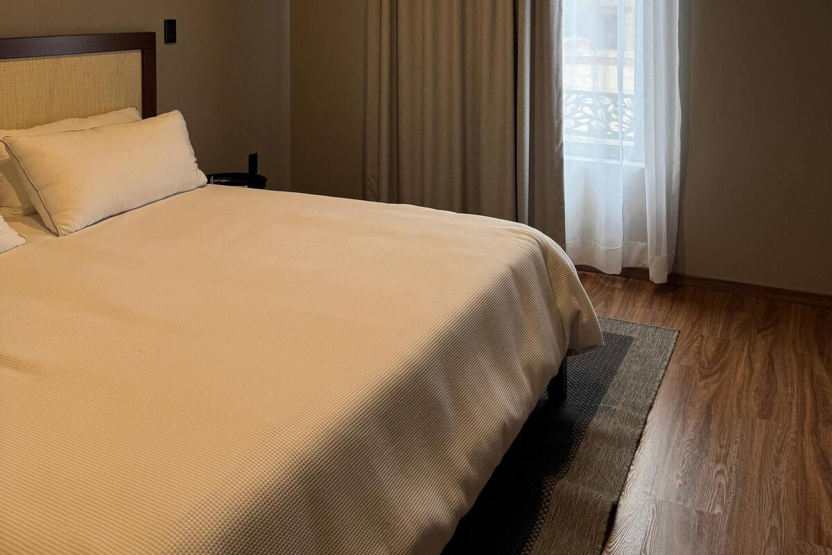 Una cama bien hecha con cabecera, mesita de noche, lámpara de pared y ventana con cortinas.