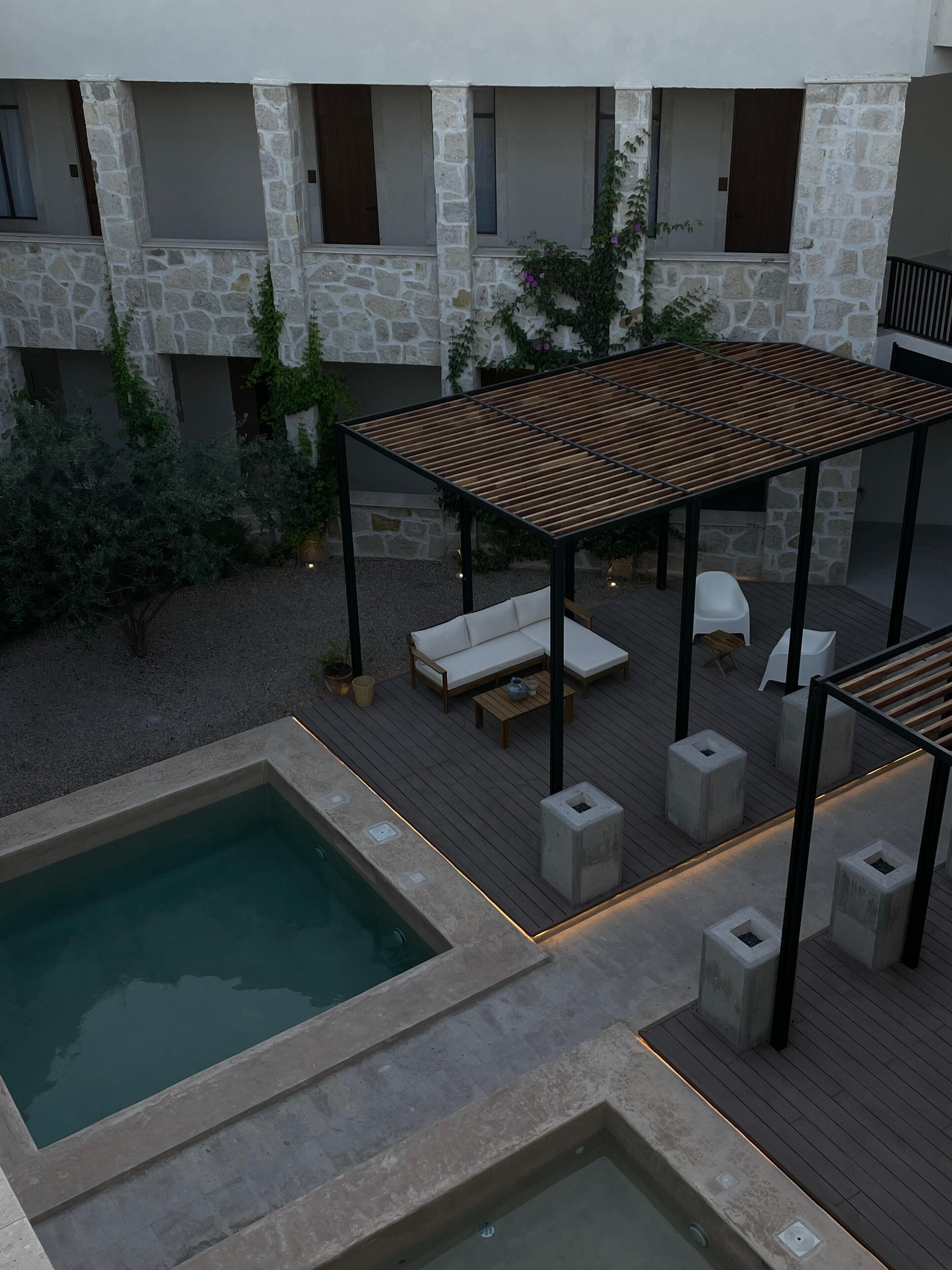 A rooftop pool area with a wooden pergola, seating, and a stone building in the background.