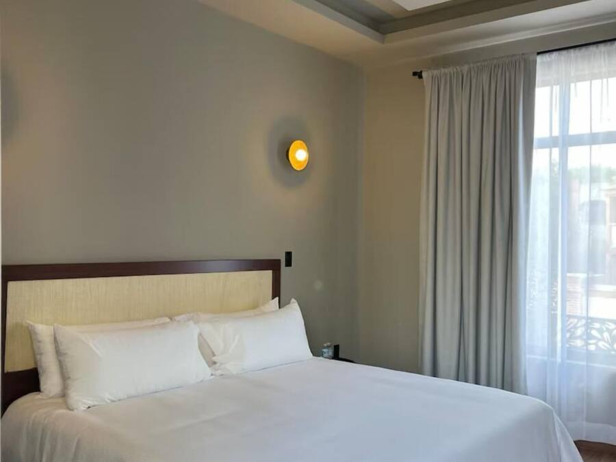 Een hotelkamer met een groot bed, hoofdeinde, een wandlamp en een raam met gordijnen.