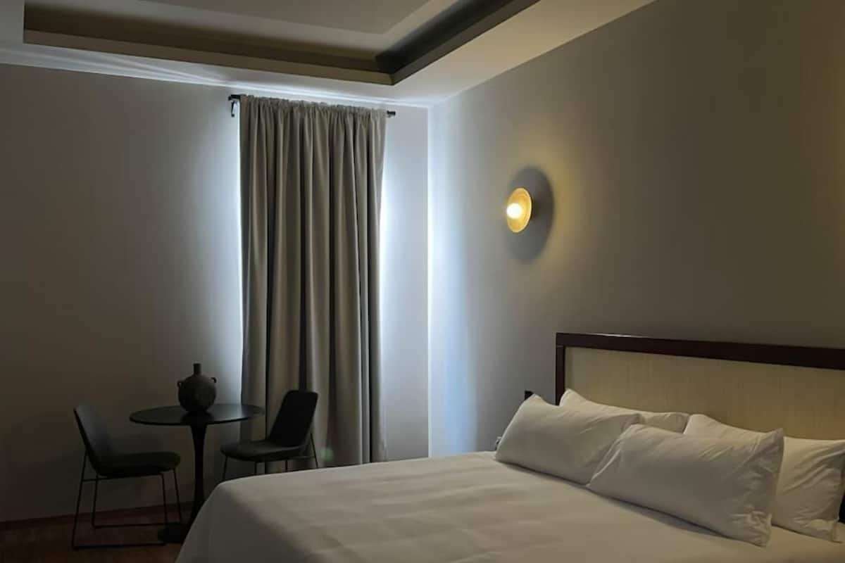 Habitación de hotel con una cama grande, una mesita con sillas, una lámpara de pared y un ventanal con cortinas.