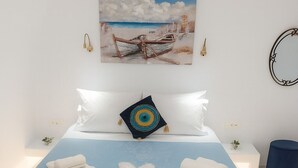 Un lit bien fait, avec des oreillers et des serviettes pliées, un tableau représentant un bateau accroché au mur et une petite plante posée sur une table de chevet.