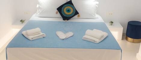 Ein ordentlich bezogenes Bett mit Kissen und gefalteten Handtüchern, ein Gemälde mit einem Boot an der Wand und eine kleine Pflanze auf einem Nachttisch.