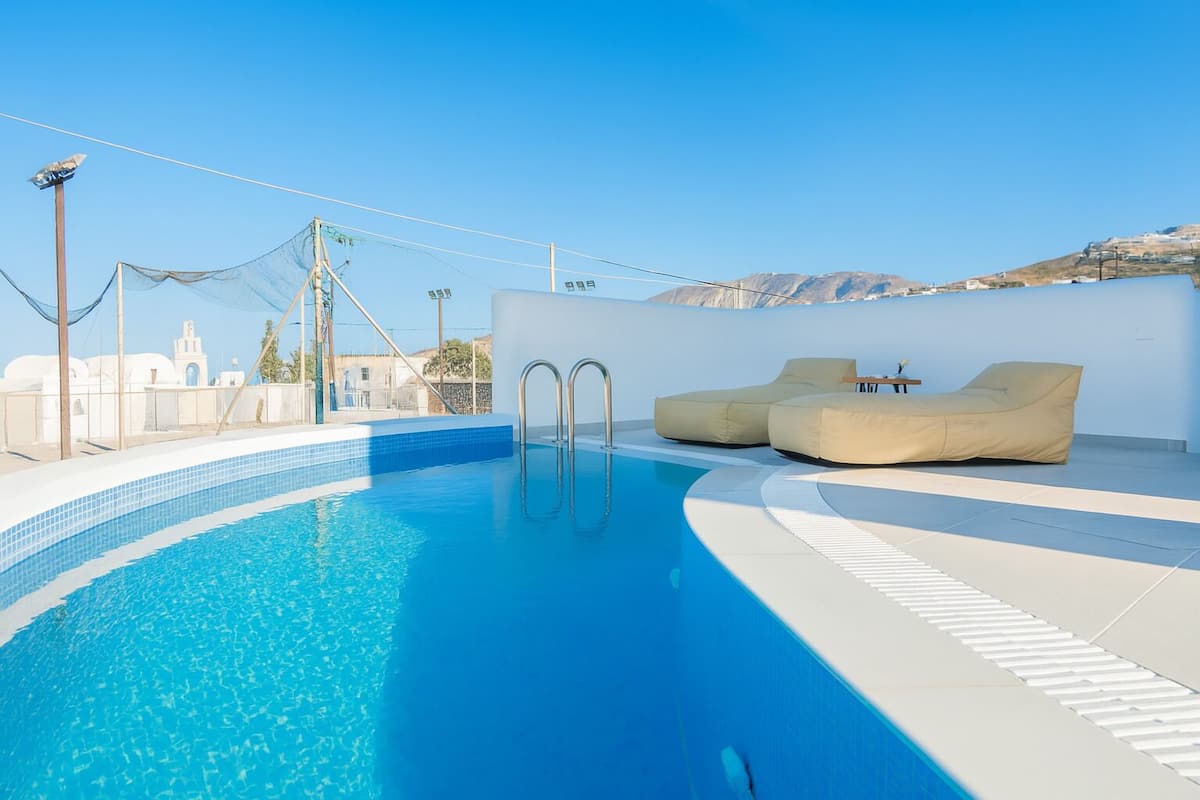 Una piscina en la azotea con dos sillones de descanso color beige y una mesa, rodeada por un muro blanco y un cielo azul despejado.