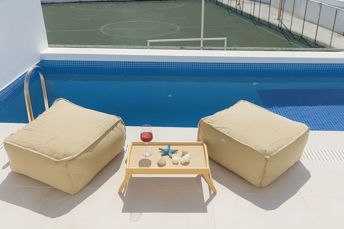 Zona junto a la piscina con dos sillas beige acolchadas, una pequeña mesa de madera con una copa de vino tinto, una estrella de mar y una concha.
