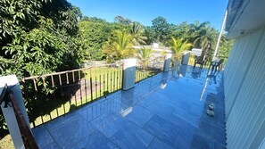 A balcony with a metal railing, a tiled floor, and a view of a lush garden with various trees and a clear sky. - A luxury escape, where the pool is always perfect (Juncos)