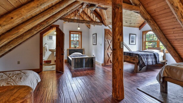 A rustic attic bedroom with exposed wooden beams, a sloped ceiling, and a large window providing natural light.
