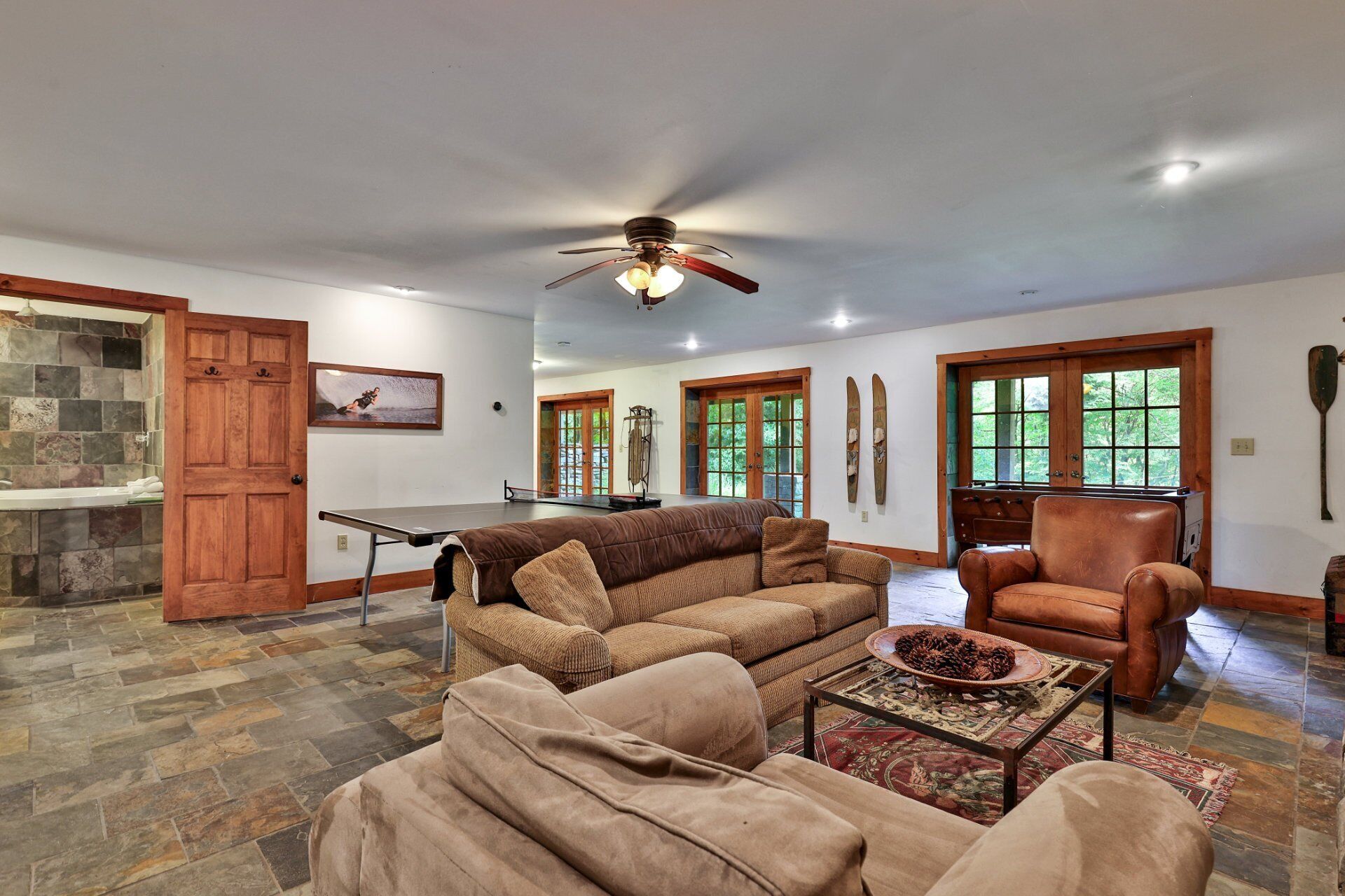 A spacious living room with a large brown sofa, a leather armchair, and a coffee table. There is a ceiling fan, a fireplace, and a stone-tiled wall.