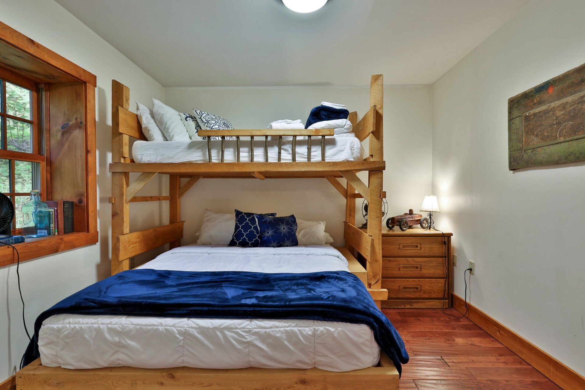 A bunk bed room with a window, a desk with a lamp, and a small shelf with books and a bottle.