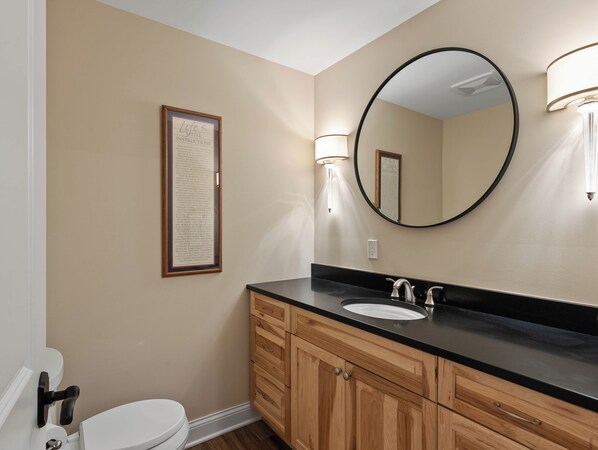 A bathroom with a white toilet, wooden cabinets, a black countertop, a round mirror, and a framed sign on the wall.