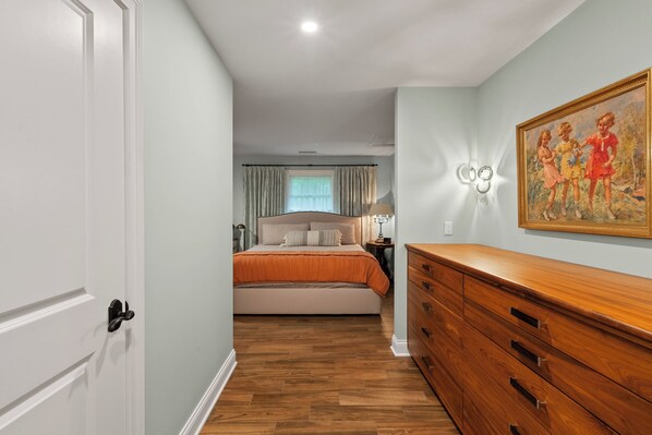 A bedroom with a wooden floor, a bed with an orange bedspread, a dresser, and a painting on the wall.