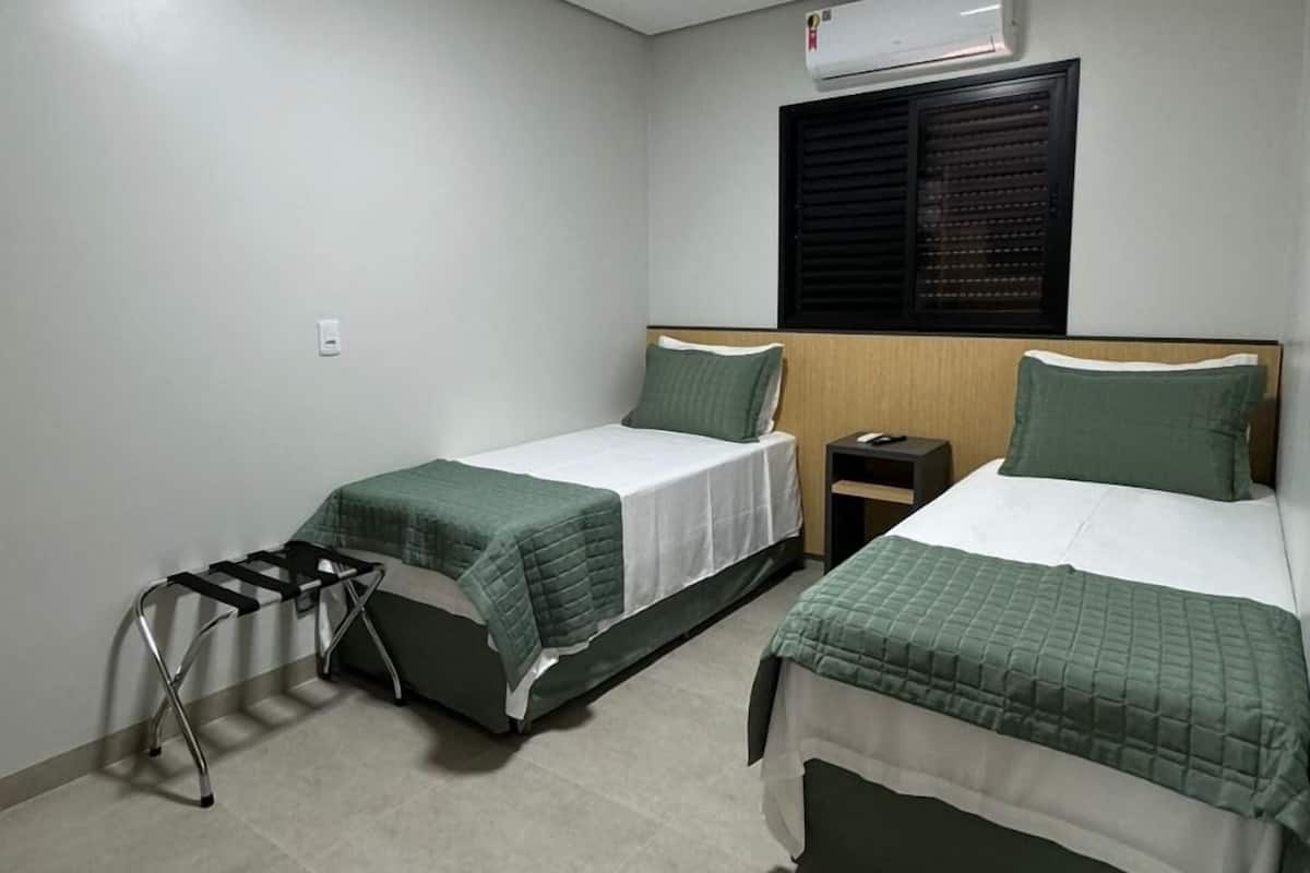 A room with two beds, a small table, a wall-mounted air conditioner, and a window with blinds.