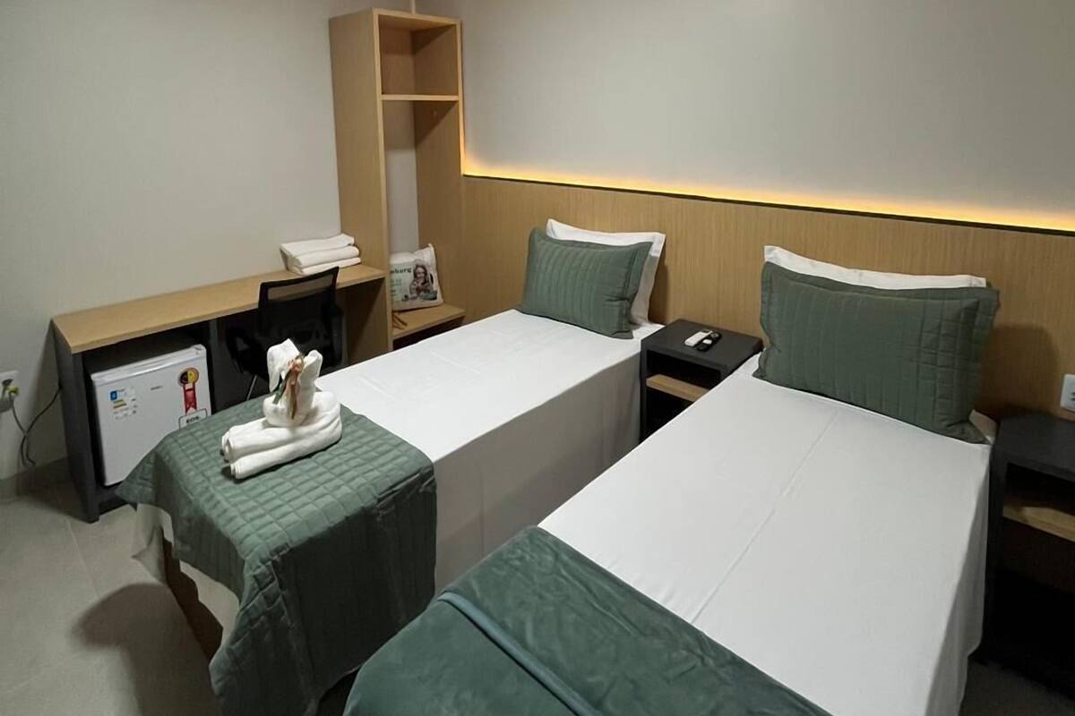 A hotel room with two single beds, a desk, a chair, a small refrigerator, and a bedside table with a phone.