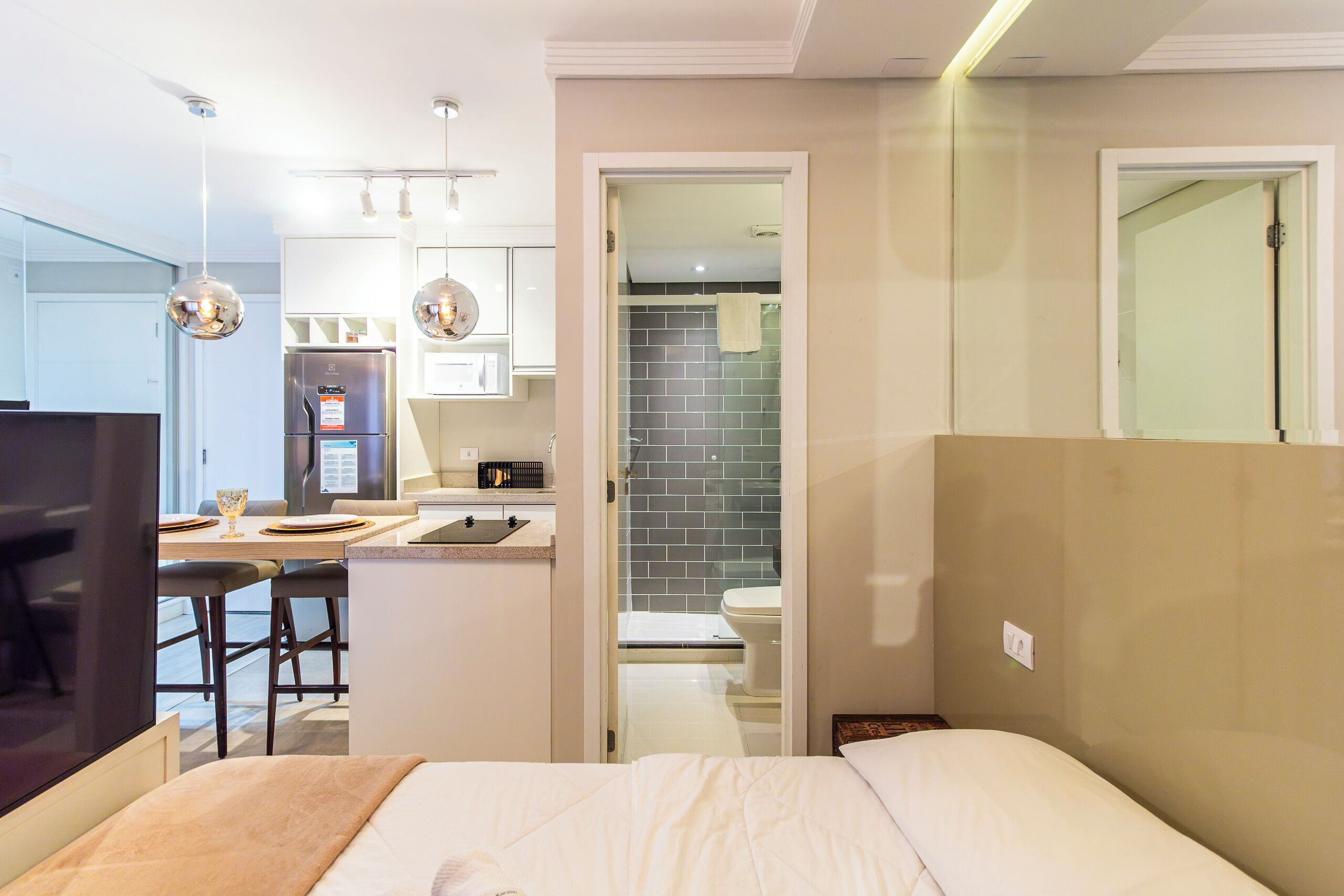 A modern hotel room with a bed, a desk with a computer, a small kitchenette, and a bathroom with a shower and toilet.