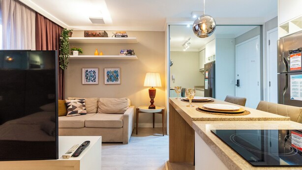 Uma sala de estar moderna com sofá, televisão e controle remoto. Inclui uma área de refeições com mesa, cadeiras e luminária.