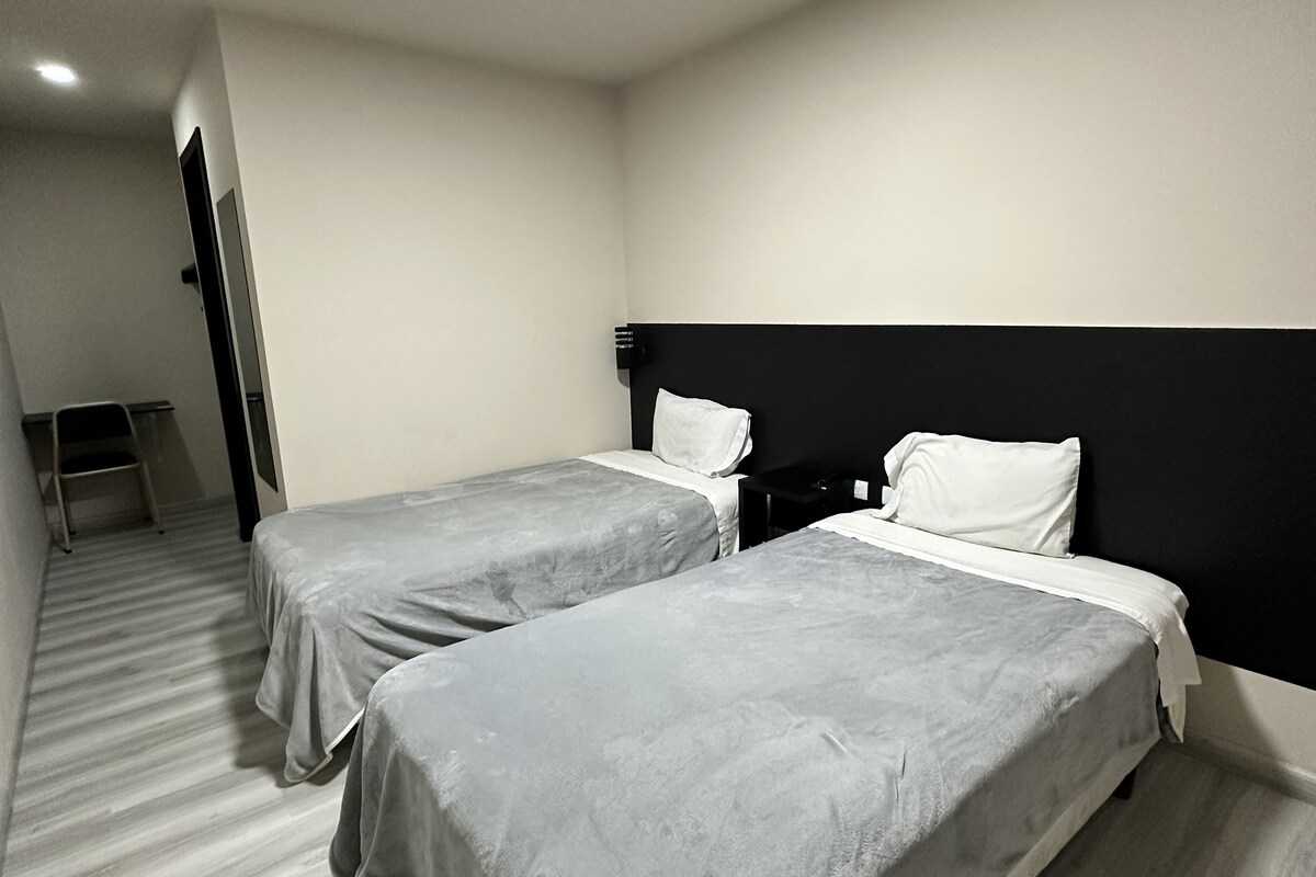 A hotel room with two single beds, a black headboard, a small desk, and a chair.
