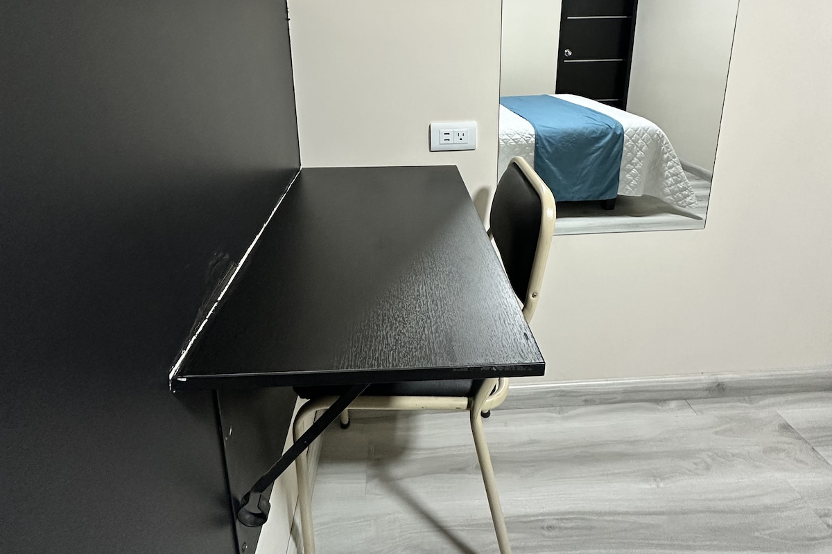 A small, modern room with a black desk and chair, a bed with a blue cover, and a mirror on the wall.