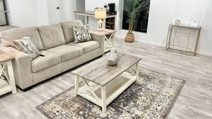 A living room with a beige sofa, a coffee table, and a patterned rug. There is a door, a plant, and a small table with a lamp. - McAllen Home Pecan - Bicentennial (McAllen)