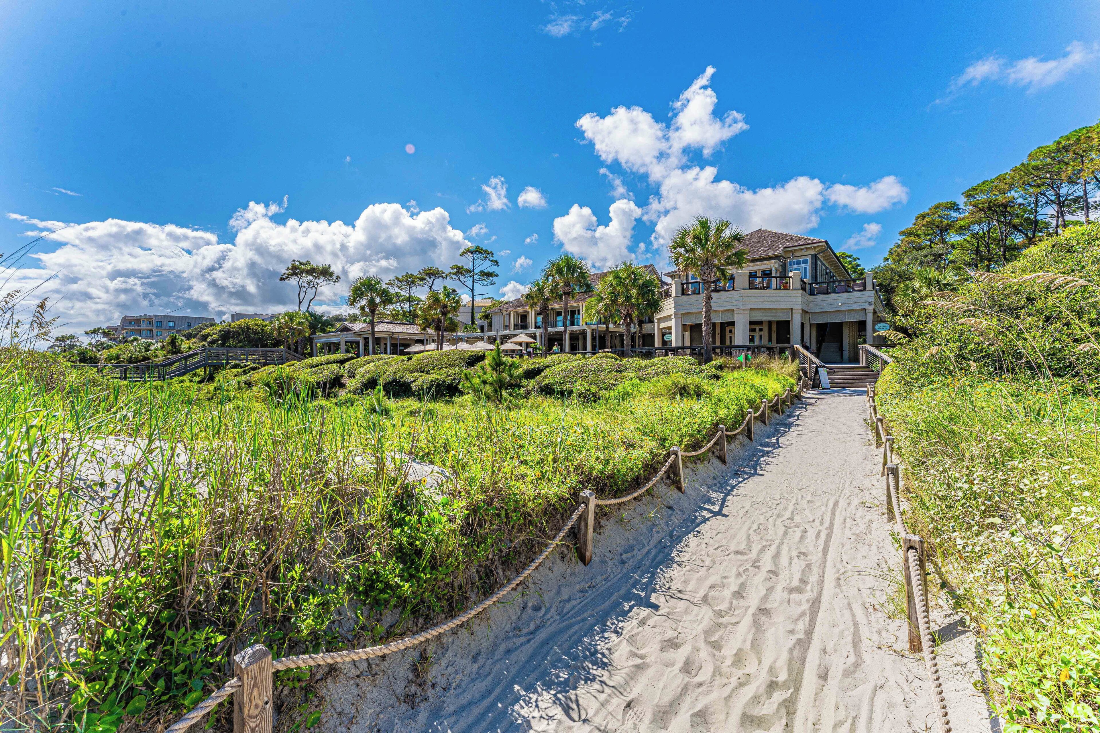 A beachfront property with a sandy pathway leading to a multi-story building, surrounded by lush greenery and palm trees.
