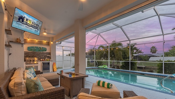 A poolside area with wicker furniture, a TV displaying 'Roelens Vacations', and a view of a swimming pool. - Canal Views, Gulf Access, Heated Saltwater Pool, Bikes - Coastal Serenity - Roelens (Cape Coral)