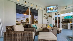 An outdoor seating area with wicker furniture, a TV screen advertising Roelens Vacations, and a barbecue grill. - Canal Views, Gulf Access, Heated Saltwater Pool, Bikes - Coastal Serenity - Roelens (Cape Coral)