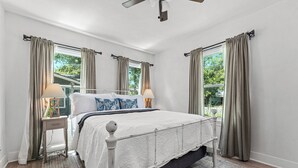 Un dormitorio con una cama, dos mesitas de noche, un ventilador de techo y ventanas con cortinas.