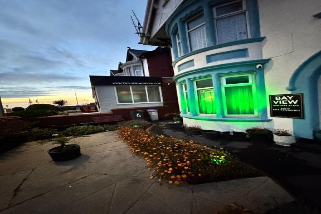 A building with a bay window illuminated by green lights, a sign reading "BAY VIEW," and a small garden with orange flowers.