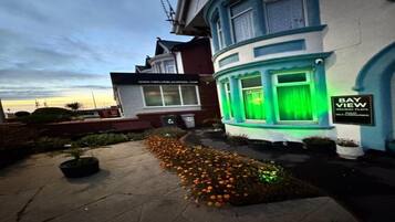 A building with a bay window illuminated by green lights, a sign reading "BAY VIEW," and a small garden with orange flowers.