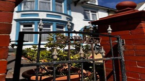 Una reja de hierro forjado con un remate decorativo, que conduce a un muro de ladrillo y una casa azul con ventanal saliente.