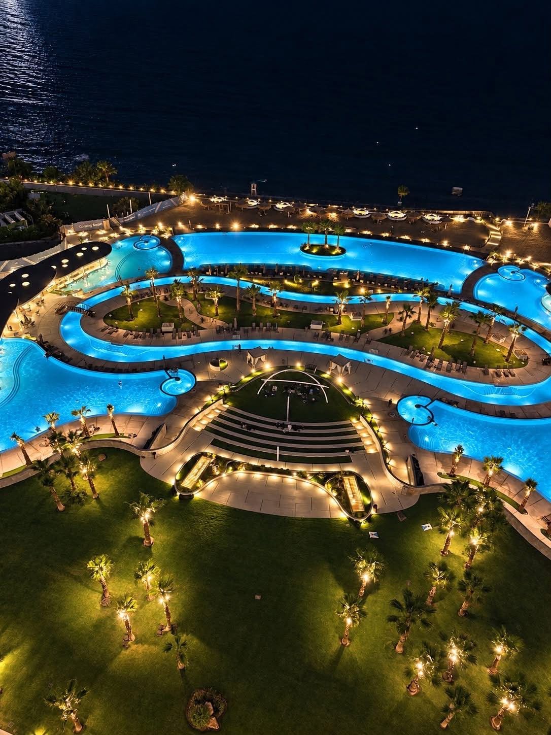 A large, multi-level swimming pool complex with surrounding lounge areas and palm trees.
