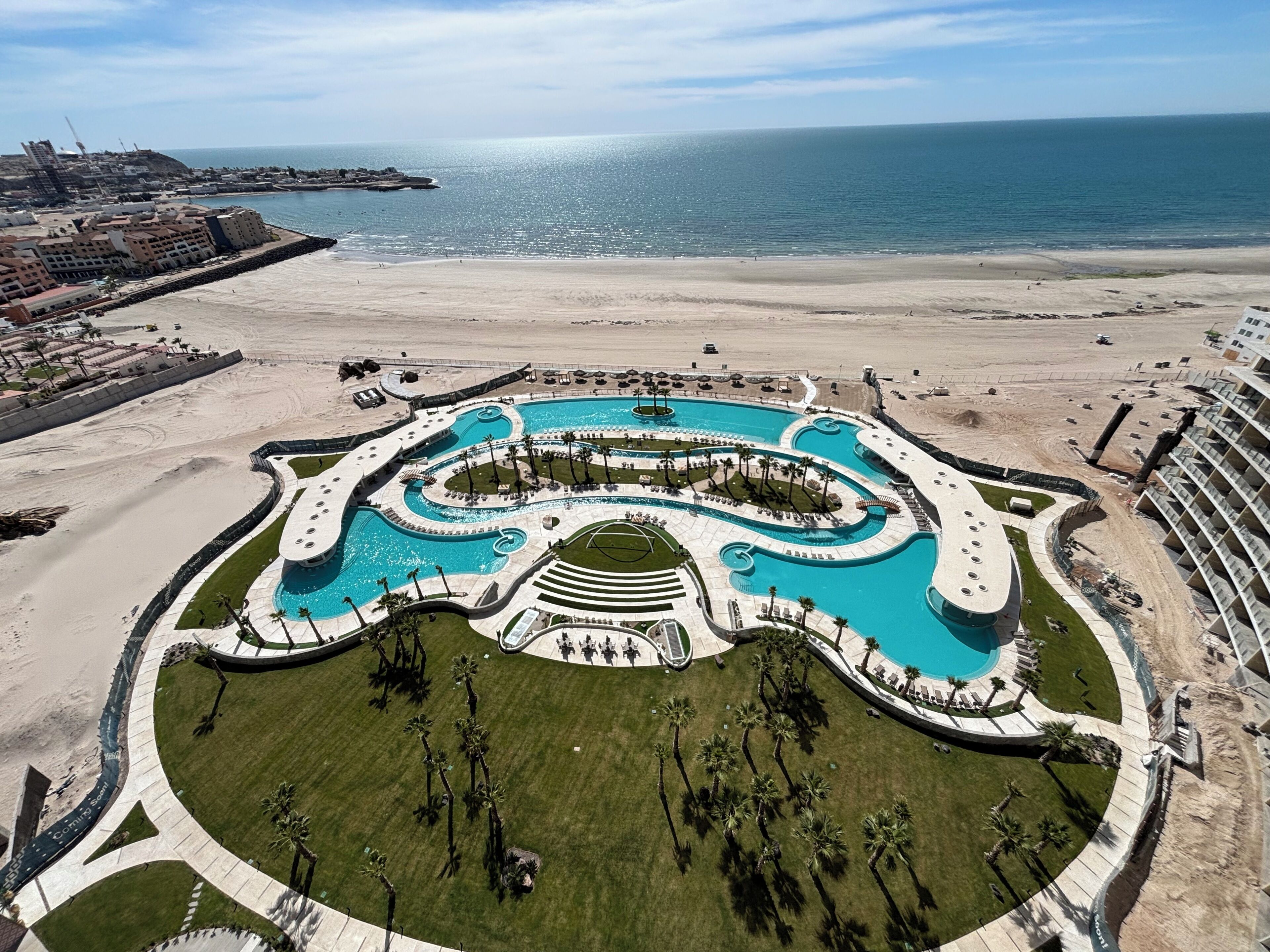 A resort with a large central pool, a sandy beach, and a view of the ocean.