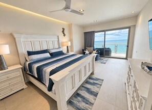 Ein Schlafzimmer mit einem großen Bett, einem Deckenventilator, einem Nachttisch, einer Kommode und Blick auf den Ozean durch eine Schiebetür.
