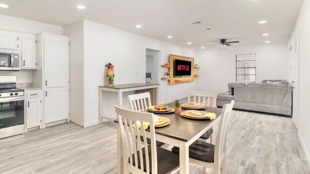 Une cuisine moderne avec des meubles blancs, un coin repas avec une table et des chaises en bois, un canapé gris et un téléviseur fixé au mur qui affiche le logo de Netflix.