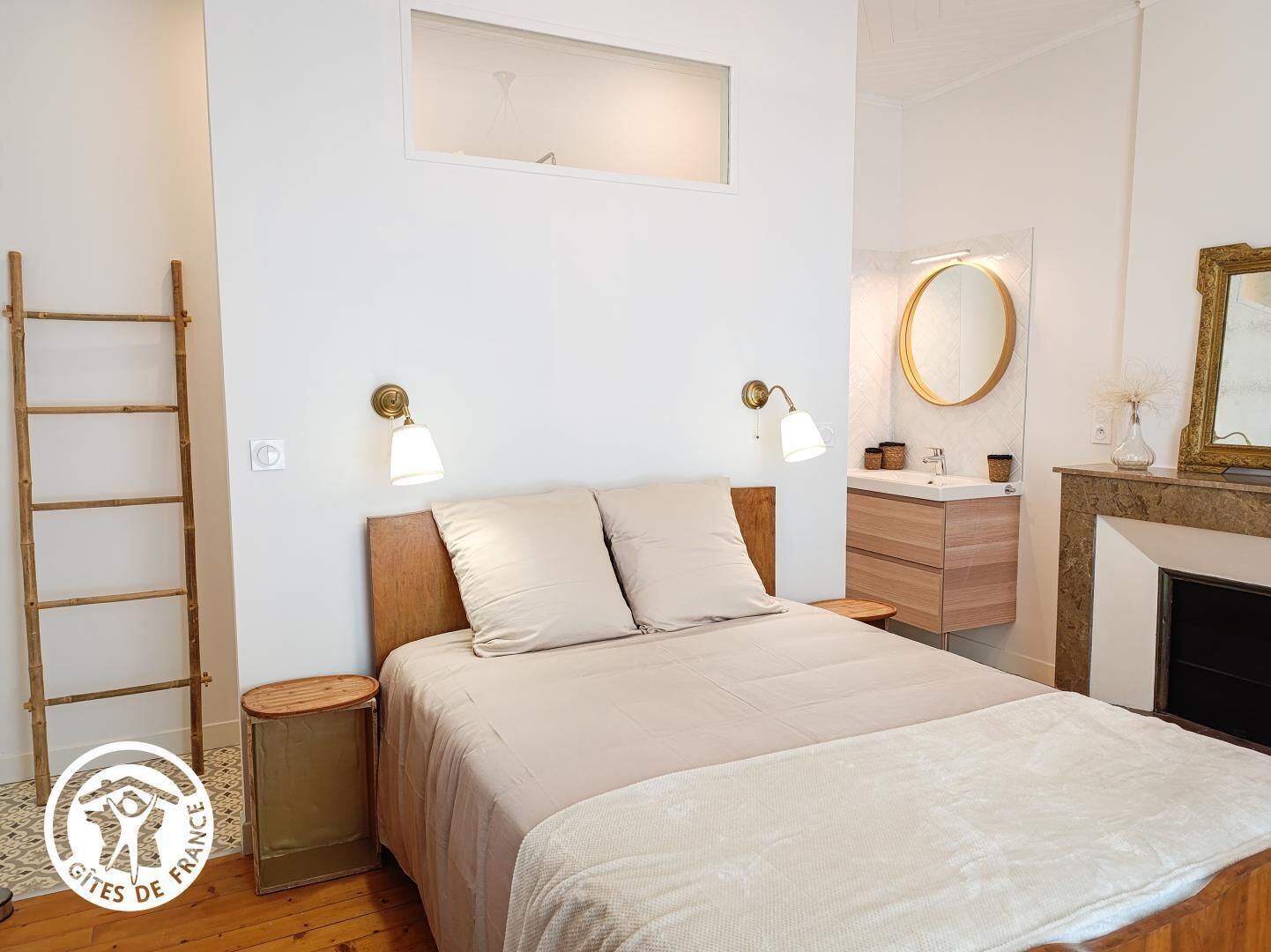 A bedroom with a wooden bed, two bedside lamps, a small round table, a mirror, and a fireplace.