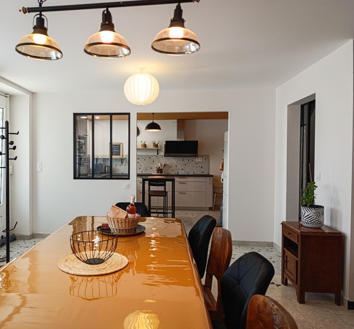 A spacious dining area with a long table, three pendant lights, and a view into the kitchen.