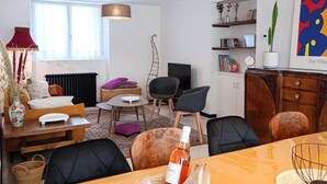Ein modernes Wohnzimmer mit einer Couch, Sesseln, einem Esstisch und einem Fernseher. Ein Bücherregal und ein Fenster mit Vorhängen sind ebenfalls vorhanden.