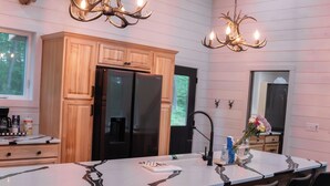 A modern kitchen with a large island, black chairs, wooden cabinets, a black refrigerator, and a deer head mounted on the wall.