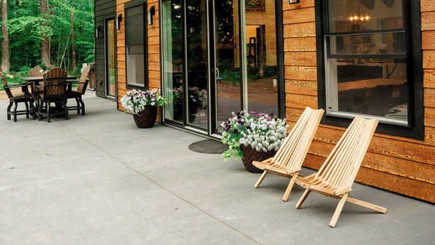 A modern two-story house with a wooden exterior, large windows, and a patio area with outdoor furniture.