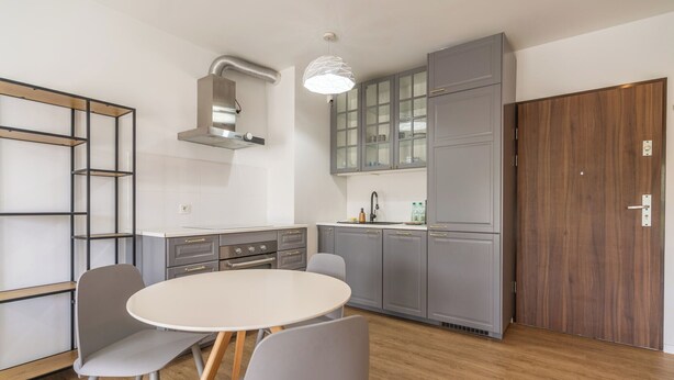 A modern kitchen with grey cabinets, a built-in oven, and a dining area with a round table and chairs. - Park Śląski Charming Apartment (Katowice)