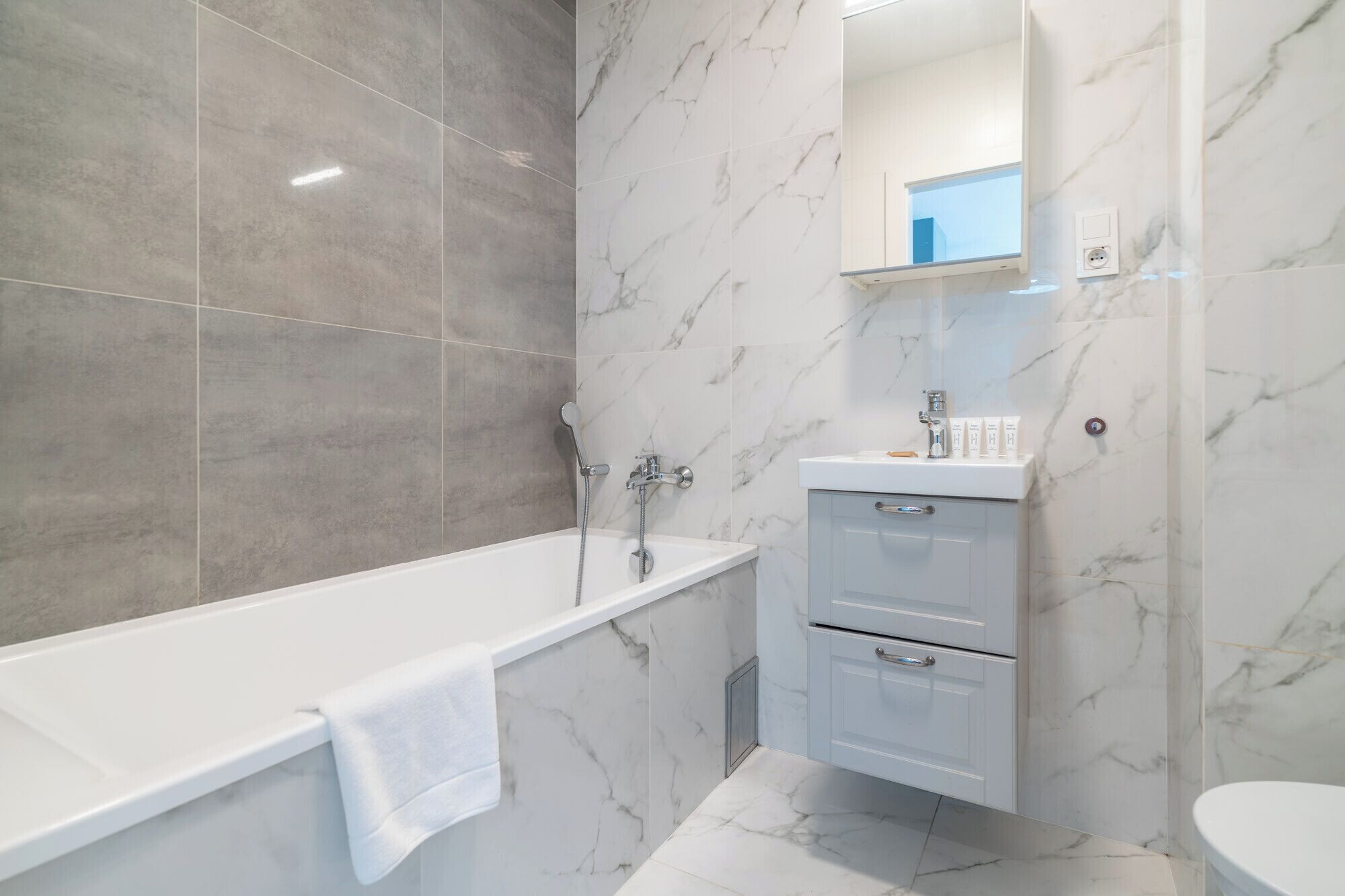 Un baño moderno con suelo y paredes de mármol, una bañera blanca, un tocador con espejo y un inodoro.