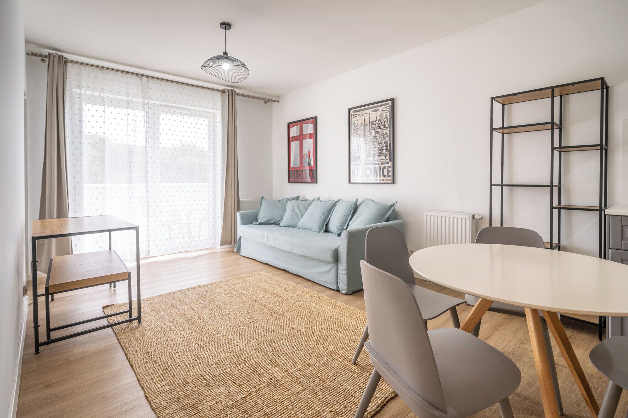 Una sala de estar moderna con un sofá, una mesa de comedor y sillas. Hay una estantería y un ventanal con cortinas.