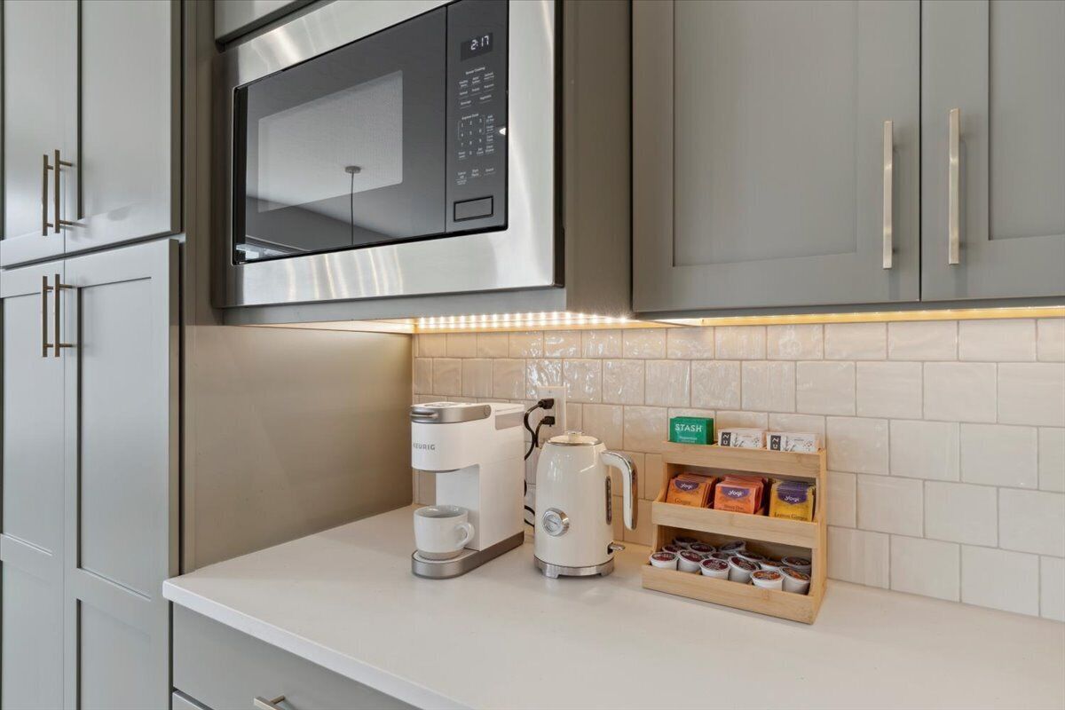 Een keuken met een roestvrijstalen magnetron, een koffiezetapparaat en een waterkoker. Er is een plank met theezakjes en een doos met de tekst 'STASH'.