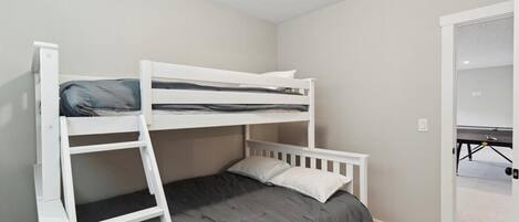 A bunk bed room with a desk and chair, a small table, and a door leading to another room.
