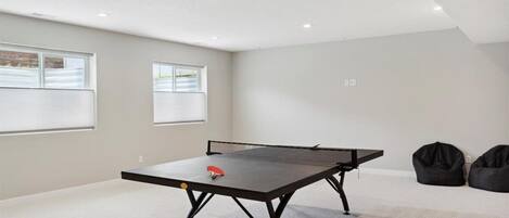 A room with a ping pong table, two windows with blinds, and two bean bags.