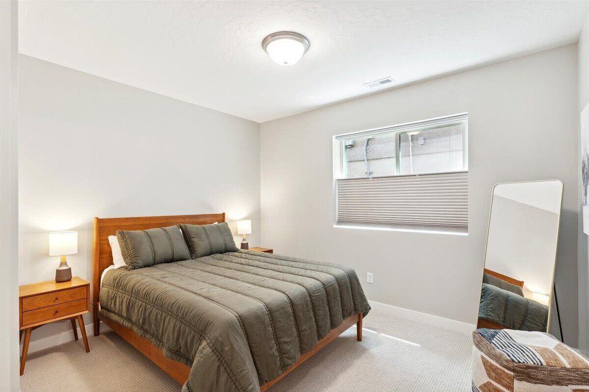 A bedroom with a wooden bed, a nightstand with a lamp, a window with blinds, and a mirror on the wall.