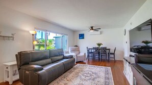 A living room with a leather sofa, a dining table with chairs, a TV, a ceiling fan, and a large window.