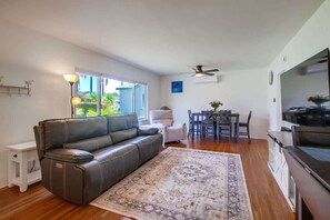 A living room with a leather sofa, a dining table with chairs, a TV, a ceiling fan, and a large window.