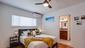 A bedroom with a bed, bedside table, ceiling fan, window, and a view into the bathroom.