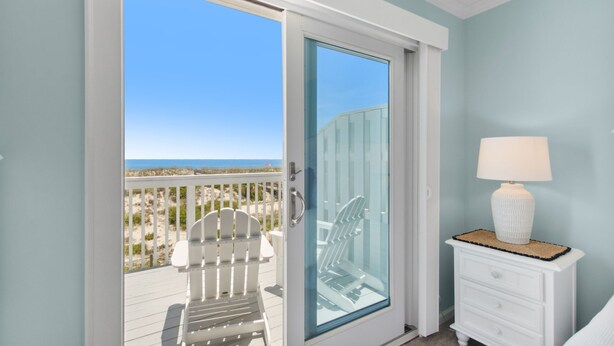 Una habitación con puerta de vidrio corredera que da a un balcón, con una silla Adirondack blanca y una mesita de noche con lámpara.