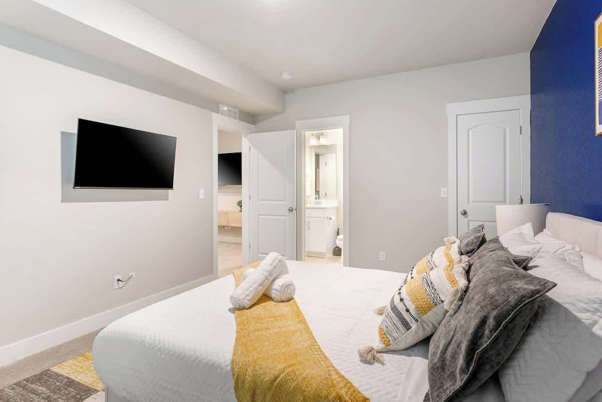 Une chambre à coucher avec un lit, un téléviseur à écran plat fixé au mur, et une salle de bain visible à travers une porte ouverte.